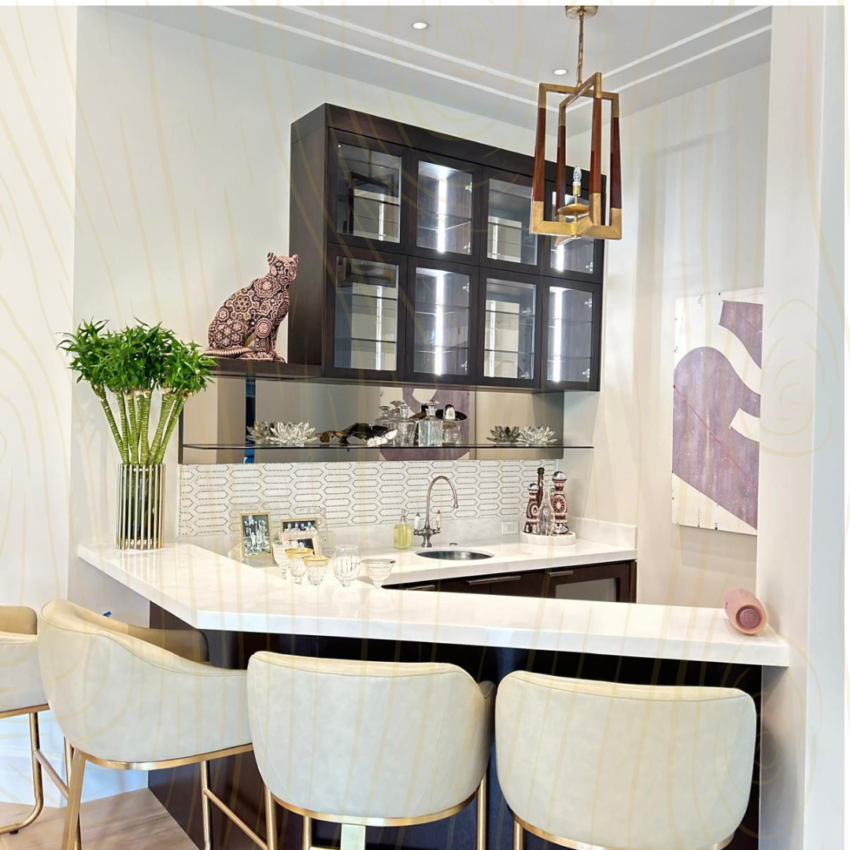 Sophisticated home bar area with white countertop, dark illuminated glass cabinets, cream leather barstools, modern chandelier, and a patterned cat statue by MR Designer Services.